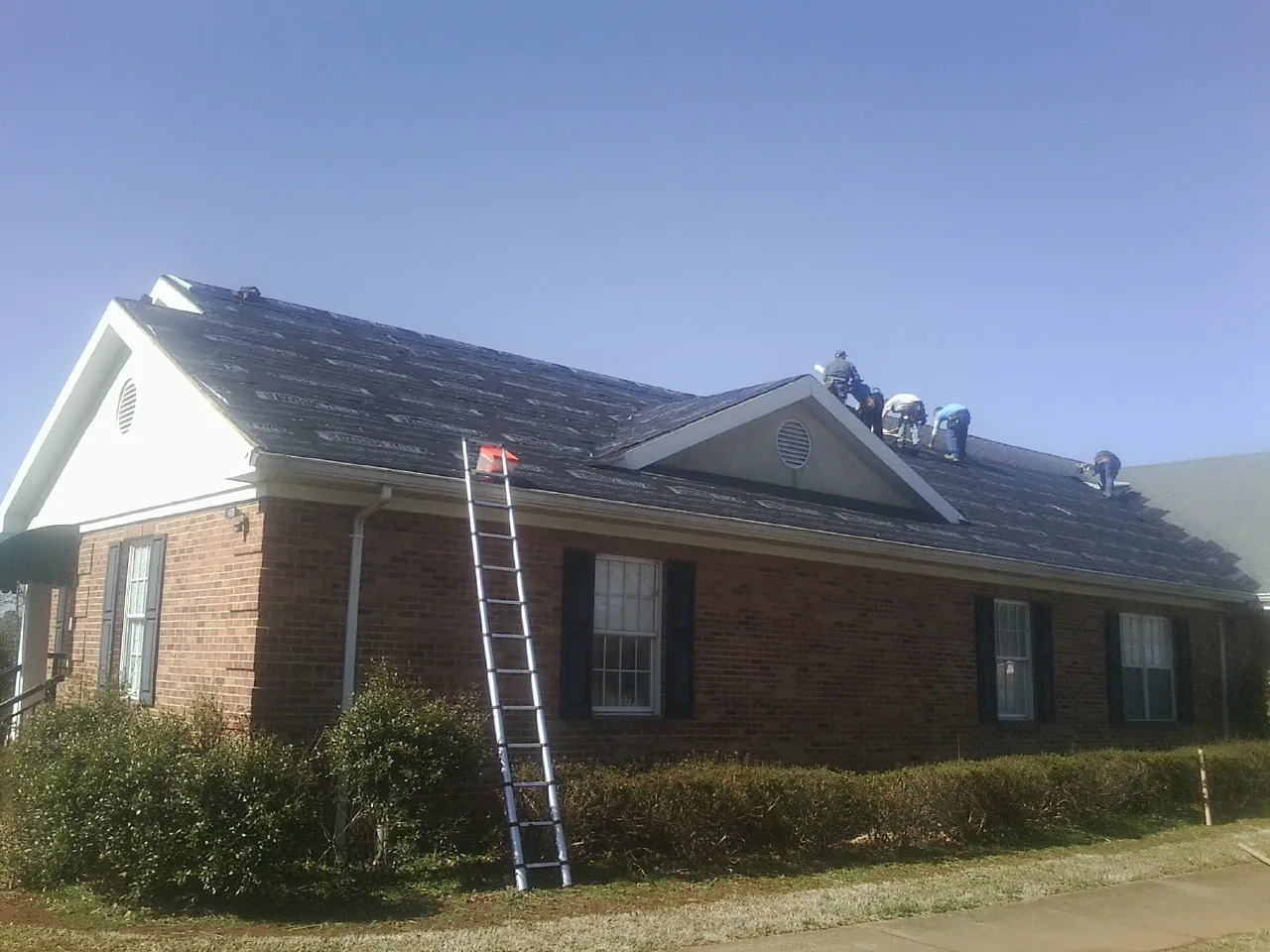Crew tearing shingles off a brick ranch roof with a ladder set — Metal Roof Repair work by our crew in Sweet Valley