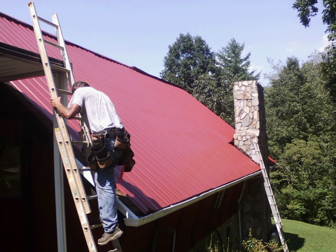 Roofer on a ladder beside a red metal roof and stone chimney — Soffit & Fascia Repair by Roofing Brothers in Sweet Valley